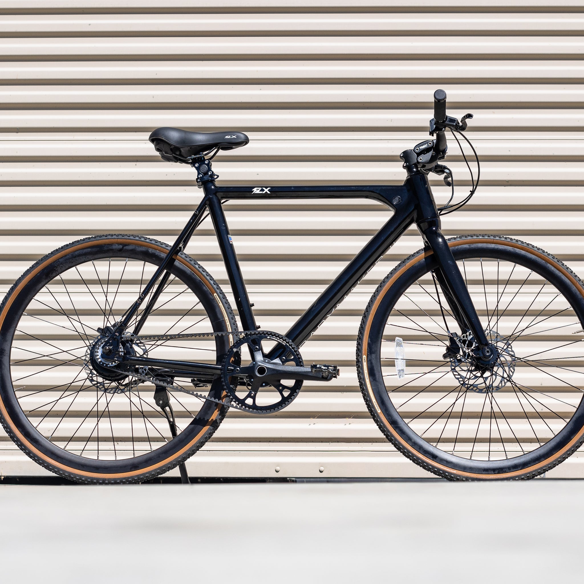 black flx babymaker II steal electric commuter bike with tan wall gravel tires and a suspension stem in front of a tan garage door