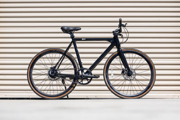 black flx babymaker II steal electric commuter bike with tan wall gravel tires and a suspension stem in front of a tan garage door