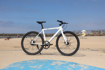 A white stealth electric commuter ebike, the flx babymaker ii on the beach in San Diego in front of a lifeguard tower